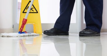 Low angle closeup of a person cleaning the floor with a mop near a yellow caution wet floor sign FLOORS & SURFACES Cleaning